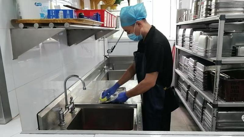 CleanLee staff in a hairnet, mask and gloves washing kitchenware at a stainless steel sink beside racks of gastronorm pans