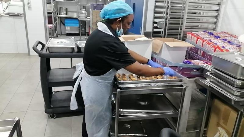 CleanLee kitchen helper in a hairnet and gloves arranging pastries on baking trays on a kitchen trolley