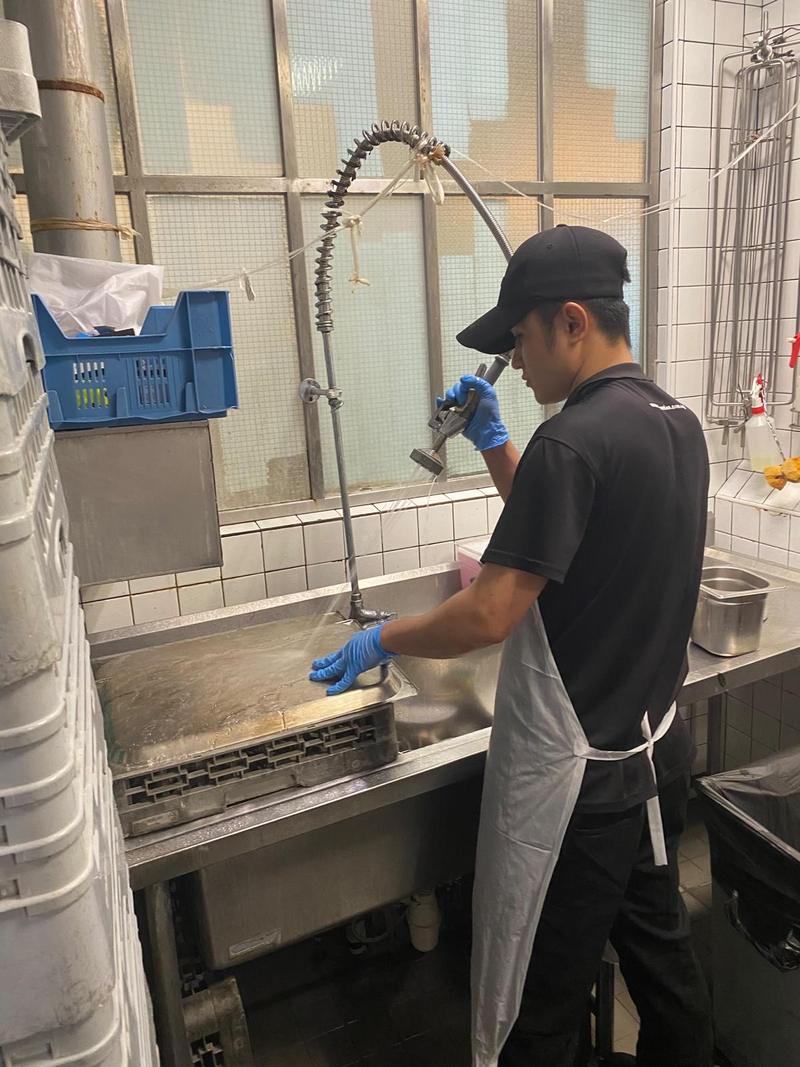 Dishwasher in black uniform, apron and gloves spraying trays at a commercial kitchen sink