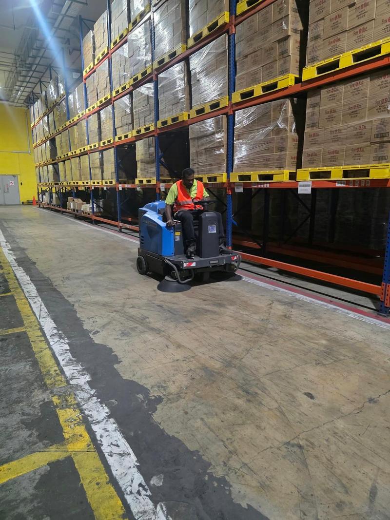 CleanLee operator driving a ride-on sweeper along a warehouse aisle lined with pallet racking
