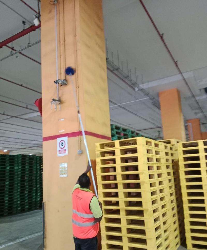 Cleaner using an extendable pole to dust a pillar beside stacked pallets in a warehouse